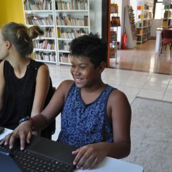 EduMais Summer Camp Program student Eryc smiling in front of a laptop in the Solar Menions de Luz library