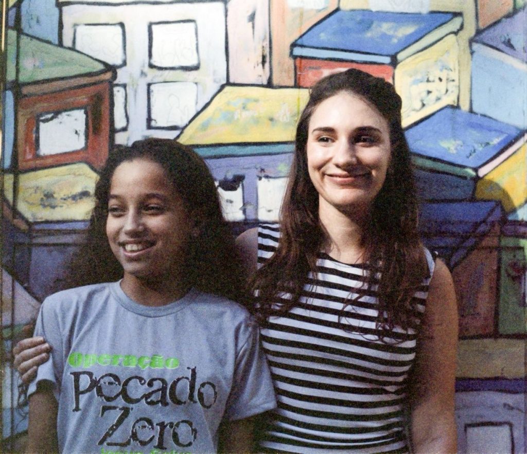 Volunteer English teacher Nikola with arm round one of her students Izabely in front a colourful favela mural