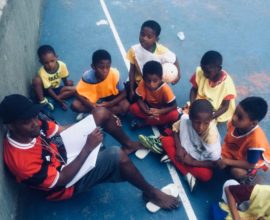 EduMais Football Program leader William sits on the soccer pitch and takes the register as the kids arrive to train