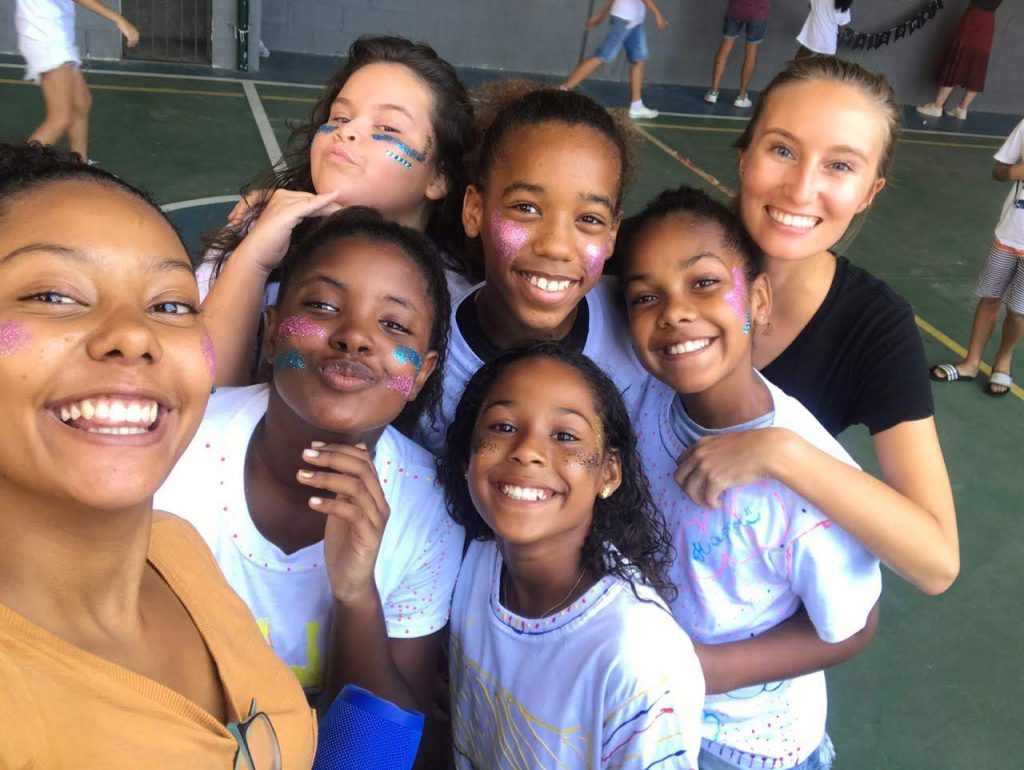 5 girls at EduMais's Summer Camp 2019 and volunteers Thais and Audrey take a selfie with their cheeks covered in glitter