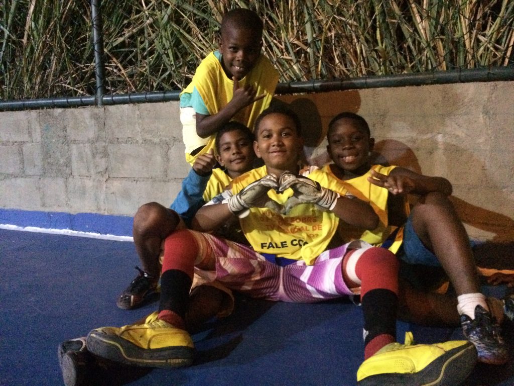 Boys on EduMais's football program sit by the wall on the pitch and the boy in the middle makes a heart shape with his hands