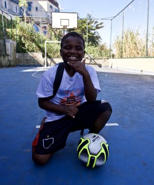 Student on EduMais's Education-Football Program knees on the pitch smiling with a soccer ball in front of him