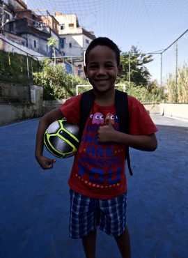 Student on EduMais's Education-Based football program holds a ball under his arm after receiving it as a donaion from Wings with favela in background