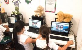 Two EduMais girl students work on their computers on the Web Design Program with cuddly toys on top of the computers