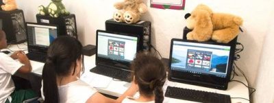 Two EduMais girl students work on their computers on the Web Design Program with cuddly toys on top of the computers