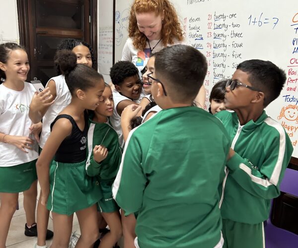 Maga li getting hugged A volunteer teacher being warmly hugged by a group of smiling children in a classroom, showing joy and connection during an EduMais lesson