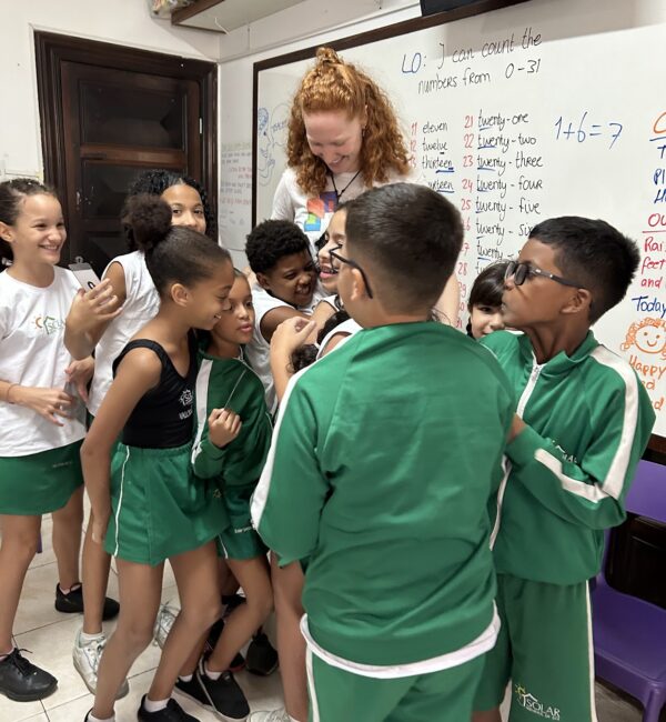 A volunteer teacher being warmly hugged by a group of smiling children in a classroom, showing joy and connection during an EduMais lesson