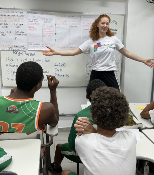 A volunteer teacher leading an English lesson with students in Rio de Janeiro, standing in front of a whiteboard with classroom guidelines and tongue twisters, engaging the class in a fun learning activity